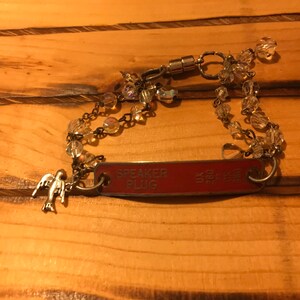 K&ouml;nnte beinhalten: Ein silbernes Armband mit einem roten Metalletikett, auf dem "SPEAKER PLUG" steht, und einem kleinen silbernen Vogelanh&auml;nger. Das Armband besteht aus klaren Glasperlen und einem Magnetverschluss.