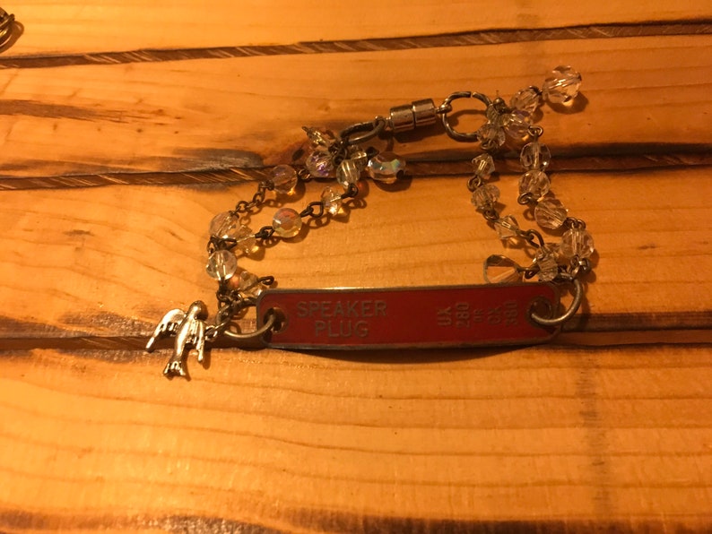 K&ouml;nnte beinhalten: Ein silbernes Armband mit einem roten Metalletikett, auf dem "SPEAKER PLUG" steht, und einem kleinen silbernen Vogelanh&auml;nger. Das Armband besteht aus klaren Glasperlen und einem Magnetverschluss.