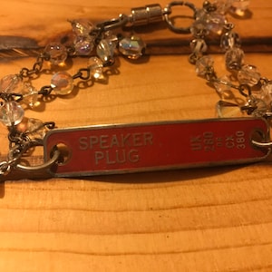 K&ouml;nnte beinhalten: Ein roter Metallanh&auml;nger mit der Aufschrift "SPEAKER PLUG", befestigt an einer silbernen Kette mit Kristallperlen und einem silbernen Vogelanh&auml;nger.