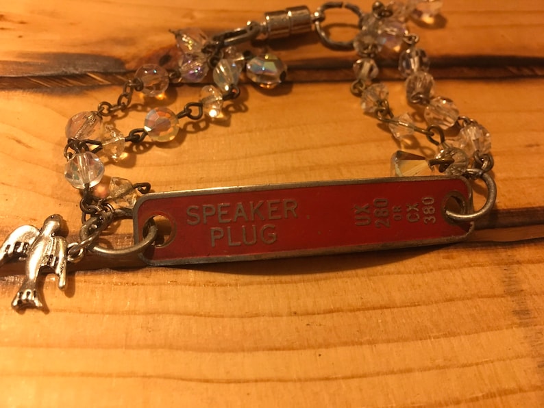 K&ouml;nnte beinhalten: Ein roter Metallanh&auml;nger mit der Aufschrift "SPEAKER PLUG", befestigt an einer silbernen Kette mit Kristallperlen und einem silbernen Vogelanh&auml;nger.