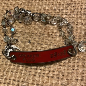 May include: A red metal bracelet with a charm that says "SPEAKER PLUG" and "UK 280 3x250". The bracelet is adorned with clear crystal beads and a silver clasp.
