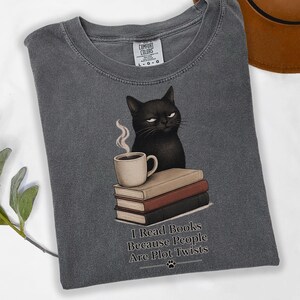 May include: A charcoal gray t-shirt featuring a graphic of a black cat sitting behind a stack of three books and a steaming mug. The text below reads, I Read Books Because People Are Plot Twists, accompanied by a small paw print icon at the bottom.