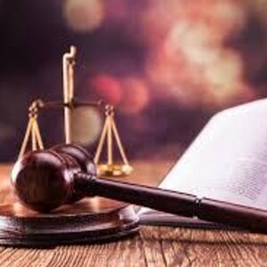May include: A wooden gavel on a round base, next to a set of golden scales and an open book. The gavel is dark brown, and the scales are balanced. The book's pages are white, and the background is blurred.