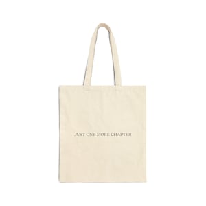 Può includere: Una borsa tote in tela color crema con due tracolle. La parte anteriore della borsa presenta il testo "JUST ONE MORE CHAPTER" stampato in un carattere semplice, di colore scuro e senza grazie, centrato al centro del corpo in tessuto.