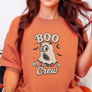 May include: A burnt orange t-shirt featuring a graphic of an owl wearing a ghost costume with the text "BOO Crew" above and below the owl, surrounded by small illustrations of bats, a crescent moon, a pumpkin, and a skull.