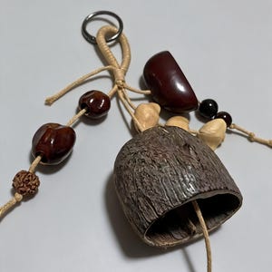 May include: A handcrafted wind chime featuring a bell-shaped object with a textured, brown surface. The chime includes various natural elements like dark brown beads, light-colored seeds, and a dark brown, half-moon shaped piece, all strung together with natural twine.