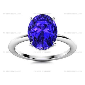 May include: A silver-toned ring featuring a large, oval-cut, vibrant blue gemstone set in a four-prong mounting. The band is smooth and polished, with the text FM GEMS JEWELLERY repeated in the background. The design is centered and symmetrical.