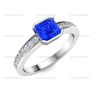 May include: A silver-toned ring featuring a square-cut, vibrant blue center stone set in a bezel setting. The band is accented with a row of small, clear, round-cut stones along each side. The text FM GEMS JEWELLERY is visible in the background.