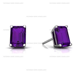 May include: A pair of stud earrings featuring rectangular emerald-cut purple amethyst gemstones set in four-prong silver-toned metal settings. The text FATIMAH GEMS JEWELLERY is repeated in the background. The earrings have a post back closure.
