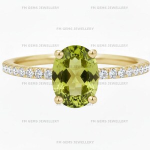 May include: A gold ring featuring a large, oval-cut, light green gemstone as the centerpiece. The band is adorned with small, clear gemstones. The words "FM GEMS JEWELLERY" are printed around the ring.