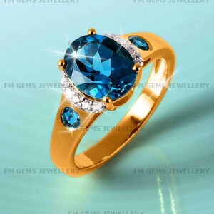 May include: A gold ring featuring a large oval-cut blue topaz center stone, accented by a halo of small white diamonds. The band includes two additional round blue topaz stones set into the gold shank on either side of the central setting.