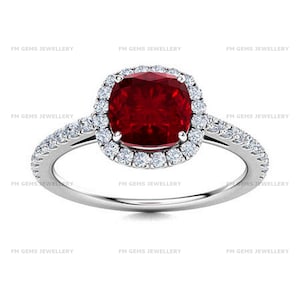 May include: A silver-toned ring featuring a central cushion-cut deep red gemstone surrounded by a halo of small white stones. The band is also set with small white stones along the sides. The text FM GEMS JEWELLERY appears repeatedly.