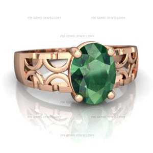 May include: Rose gold ring featuring an oval-cut, emerald-green gemstone. The band has a decorative openwork design. The ring is displayed on a reflective surface, highlighting the gemstone's facets and the band's intricate details.