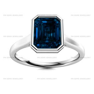 May include: A silver-toned ring featuring a large, rectangular, emerald-cut dark blue gemstone set in a smooth bezel setting. The band is polished and solid, with the text FM GEMS JEWELLERY repeated multiple times across the image background.