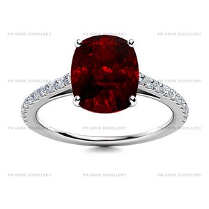 May include: A silver-toned ring featuring a large, oval-cut deep red gemstone held by four prongs. The band is accented with small, clear pavé-set stones along the shoulders. The text FM GEMS JEWELLERY is repeated across the image background.