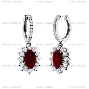 May include: A pair of silver hoop earrings featuring a dangling oval-cut deep red gemstone surrounded by a halo of small round white stones. The hoop portion of each earring is also encrusted with a row of small round white stones.