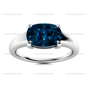 May include: A silver-toned ring featuring a large, cushion-cut, deep blue gemstone set in a four-prong mounting. The band is smooth and polished, with the text FM GEMS JEWELLERY repeated across the image background in a light gray font.