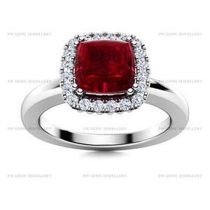 May include: A silver-toned ring featuring a central cushion-cut deep red gemstone surrounded by a halo of small, clear, round-cut stones. The band is smooth and polished, and the text FM GEMS JEWELLERY is repeated across the image background.