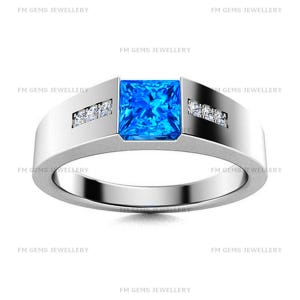 May include: A silver-toned band ring featuring a central square-cut bright blue gemstone flanked by two small clear stones on each side. The metal surface has a smooth, polished finish. The text FM GEMS JEWELLERY is repeated across the image.