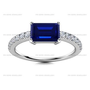 May include: A silver-toned ring featuring a rectangular emerald-cut deep blue sapphire centre stone, flanked by a row of small round white stones along each side of the band. The text FM GEMS JEWELLERY is repeated across the image background.
