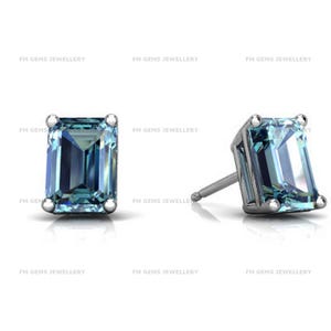 May include: A pair of stud earrings featuring emerald-cut light blue gemstones set in silver-toned metal with four prongs. The text FM GEMS JEWELLERY is repeated across the image. Each earring has a post back for secure attachment to the earlobe.