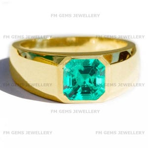 May include: A gold ring featuring a large, emerald-cut, bright green gemstone. The ring has a wide, polished gold band. The gemstone is set in a square bezel setting. The ring is displayed on a white background.