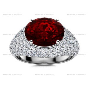 May include: A silver ring featuring a large, oval-cut deep red gemstone held by four prongs. The wide band is densely encrusted with numerous small, clear, round-cut stones. The text FM GEMS JEWELLERY is repeated across the image background.