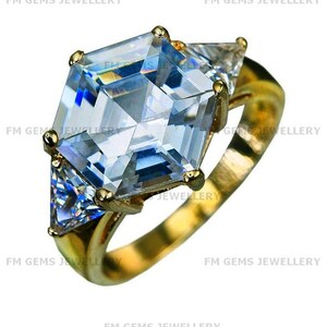May include: A gold ring featuring a large, central hexagonal-cut clear gemstone flanked by two smaller triangular-cut clear gemstones. The setting is made of polished yellow gold, and the text FM GEMS JEWELLERY is overlaid on the image.
