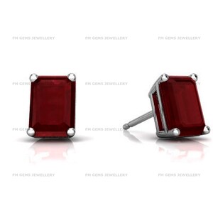May include: A pair of stud earrings featuring rectangular emerald-cut deep red gemstones set in polished silver-toned metal prongs. The design includes a post backing for secure attachment. The text FM GEMS JEWELLERY is visible in the background.