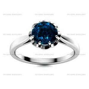 May include: A silver-toned ring featuring a round, deep blue gemstone set in a floral-shaped prong setting. The band is smooth and polished, with the text FM GEMS JEWELLERY repeated across the background. The design centers on the dark blue stone.