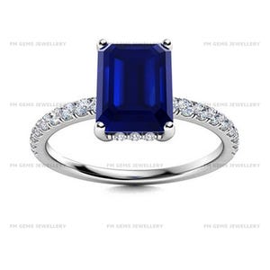 May include: A silver-toned ring featuring a large, rectangular, emerald-cut deep blue gemstone set in the centre. The band is adorned with a row of small, round, clear white stones along both sides, extending from the central setting.