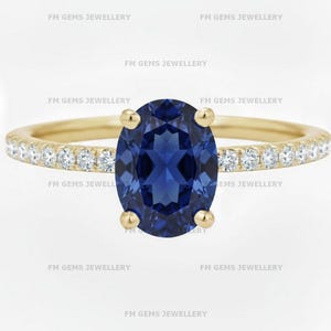 May include: A gold ring featuring a large, oval-cut, deep blue gemstone as the centerpiece. The band is adorned with small, clear gemstones. The ring is set against a white background, with the text "FM GEMS JEWELLERY" repeated around the image.
