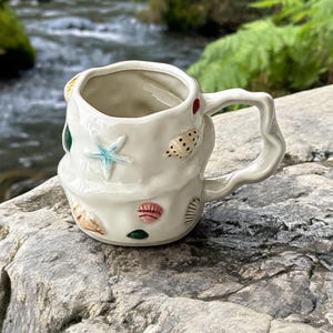 May include: Cream ceramic mug with a textured finish and a distinctive handle. The mug is decorated with seashell and starfish designs in multiple colours. The mug is placed on a rock near a river.