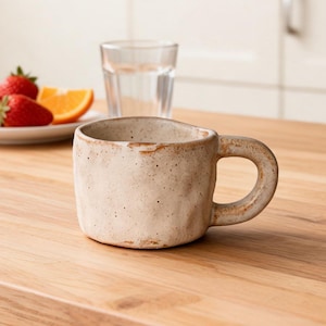 Puede incluir: Una taza de cerámica beige con un asa redondeada se encuentra sobre una superficie de madera. La taza tiene un aspecto texturizado y moteado. Un vaso de agua y un plato con fresas y rodajas de naranja están en el fondo.