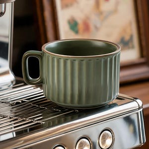 May include: A sage green ceramic coffee mug with vertical ribbed detailing and a brown rim. The mug has a curved handle and sits on a stainless steel espresso machine. The background includes a framed picture and wooden furniture.