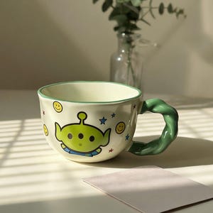 May include: A ceramic mug with a green alien character design, smiley faces, and stars. The mug has a green handle and rim, and a light beige body. The mug is on a white surface.