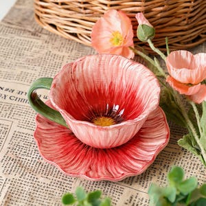 Könnte beinhalten: Ein Keramik-Tee-Set in Form einer Mohnblume. Die Tasse und Untertasse sind hauptsächlich rot mit weißen Akzenten, der Henkel ist grün. Das Set steht auf einer Zeitung, im Hintergrund ein Weidenkorb und Mohnblumen.