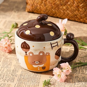 May include: A ceramic mug with a brown mushroom-shaped lid. The mug features a bear illustration wearing an orange outfit. The handle is brown, and the mug is decorated with small brown acorns and yellow dots.