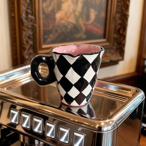 May include: A ceramic mug with a black and white checkered pattern and a pink interior. The mug has a black handle and sits on a stainless steel surface. The mug's rim has a wavy design.
