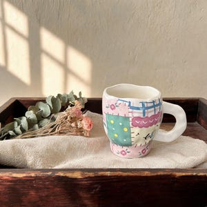 Puede incluir: Una taza de cerámica blanca con una forma irregular única y un asa grande. La taza presenta un diseño de patchwork en rosa, azul, verde y morado, con motivos florales y geométricos. La taza está colocada sobre una bandeja de madera con flores secas.
