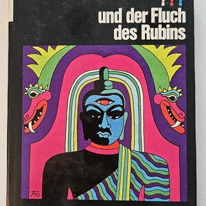 Könnte beinhalten: Ein gebundenes Buch mit dem Titel Alfred Hitchcock Die drei ??? und der Fluch des Rubins mit schwarzem Einband und einer farbenfrohen Illustration einer blaugesichtigen Figur mit drittem Auge, flankiert von zwei stilisierten Drachen in Pink und Grün.