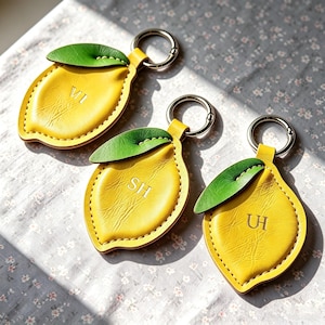 Peut inclure: Trois porte-clés en cuir en forme de citron, jaune vif, avec une feuille verte sur le dessus. Chaque porte-clés possède un anneau en métal argenté et est gravé de deux initiales : VI, SH et UH, respectivement, dans une police serif.