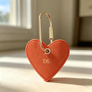 May include: A heart-shaped orange keychain with a gold-colored clasp. The keychain has the initials "DL" in gold lettering. The background is a soft, neutral color, with a window visible in the distance.