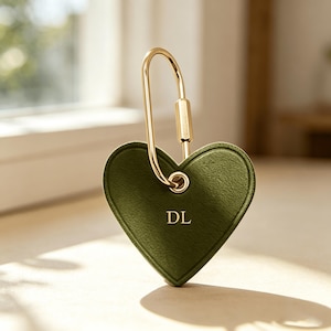 May include: A heart-shaped, olive green leather keychain with gold-colored hardware. The keychain features the initials "DL" and a gold-colored clasp. The design is simple and elegant.