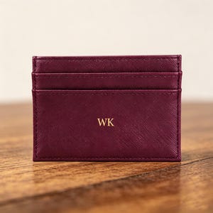 May include: A burgundy card holder with three card slots. The front of the holder has the gold letters "WK". The card holder is made of a textured material and sits on a wooden surface.