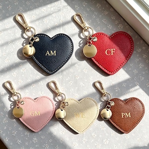 Könnte beinhalten: Fünf herzförmige Schlüsselanhänger aus lederähnlichem Material in Marineblau, Rot, Hellrosa, Creme und Braun. Jeder Anhänger hat goldfarbene Beschläge, einen Karabinerverschluss, einen kleinen runden Anhänger und goldene Initialen: AM, CF, GM, WE und PM.