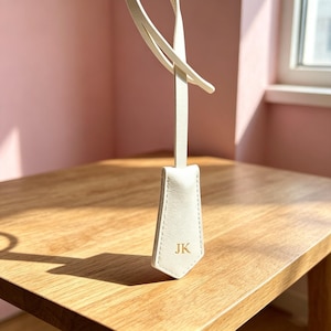 May include: A white luggage tag with a long, thin strap and a pointed bottom edge. The tag features the gold-colored embossed initials JK centered on the front surface. The design includes visible stitching along the edges of the tag body.