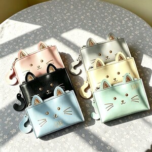 May include: A collection of six rectangular coin purses in pastel and black colours, each featuring cat ears with gold glitter accents, a cat face print with whiskers, and a curved tail-shaped zipper pull. Two purses display the letters QE and BP.