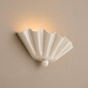 May include: A wall-mounted light fitting featuring a glossy, off-white ceramic shell-shaped shade with vertical fluted pleats. The design includes a small, spherical accent knob at the base, emitting a soft, warm glow from the top opening.