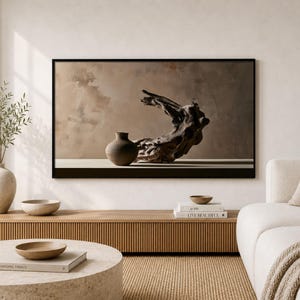 Könnte beinhalten: Ein rechteckiges Wandbild in einem schwarzen Rahmen, das eine dunkelbraune Keramikvase und ein Stück verwittertes Treibholz vor einem beigen Hintergrund zeigt. Das Kunstwerk hängt über einer hölzernen Konsole in einem hellen Wohnzimmer.
